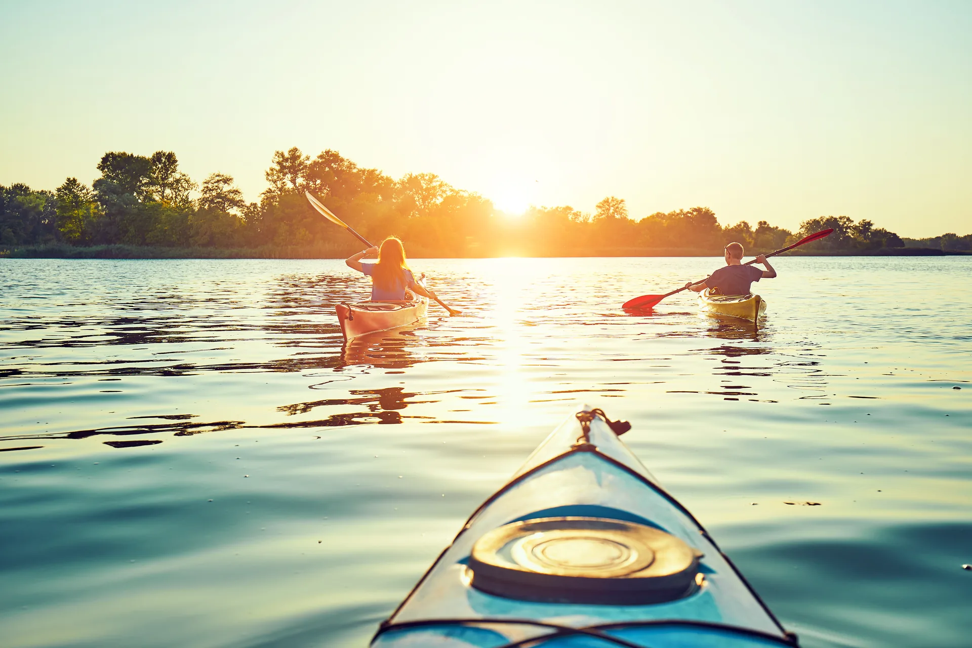 Two kayakers paddling toward a low sun on a calm lake, seen from the bow of a third kayak